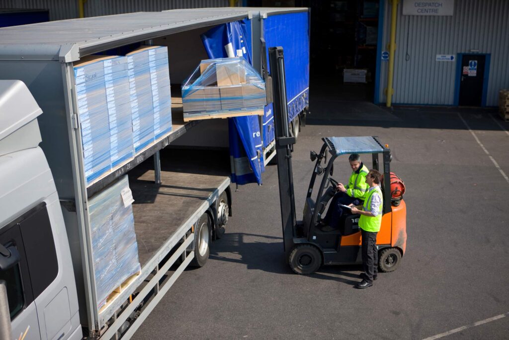 A middle aged worker driving and using the forklift to load packages on a semi-truck outside the warehouse with the help of a colleague standing beside