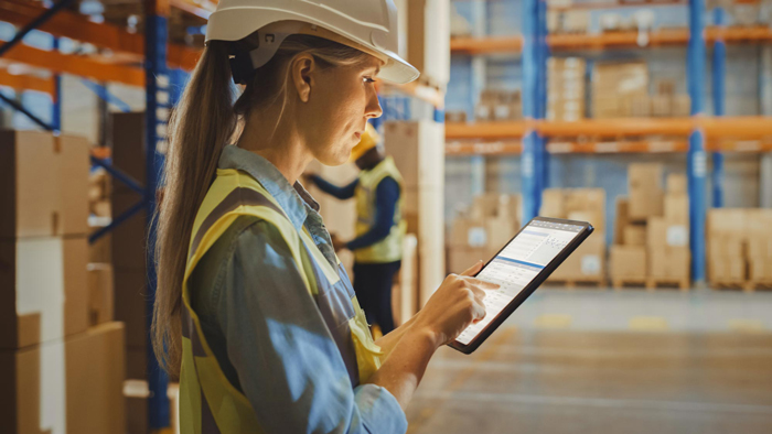 Professional Female Worker Wearing Hard Hat Uses Digital Tablet Computer with Inventory Checking Software in the Retail Warehouse full of Shelves with Goods